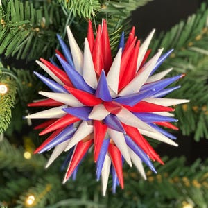May include: A festive Christmas tree ornament with a starburst design. The ornament features alternating red, white, and blue pointed shapes, creating a patriotic color scheme. The ornament is displayed on a green Christmas tree.