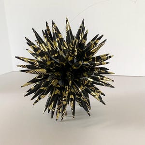 May include: A decorative, spiky ornament with a spherical shape. The ornament features a black base with gold metallic accents, creating a striking contrast. The spiky elements radiate outwards, giving it a starburst appearance. A thin, clear string is attached for hanging.