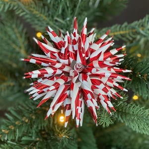 May include: A red and white striped starburst Christmas ornament. The ornament is made of metallic material and has a spiky design. It is hanging on a green Christmas tree with small yellow lights.