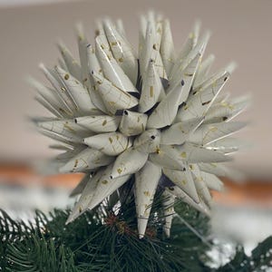 May include: A white and gold star-shaped Christmas tree topper. The star has many cone-shaped points with gold accents. The topper is placed on top of a green Christmas tree.