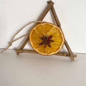 Może przedstawiać: Suszony plaster pomarańczy z gwiazdą anyżu w środku, zawieszony w trójkątnej ramie z gałązek. Do zawieszenia przymocowana jest pętla z naturalnego sznurka. Plaster pomarańczy ma średnicę około 7,6 cm.