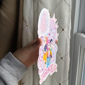 May include: A pink and white birthday cake topper with the number 6, musical notes, and stars. The topper features a group of cartoon characters and the name "GRACE" in a banner. The design is held by a person.