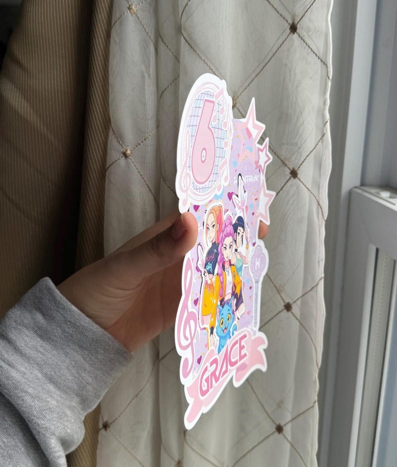 May include: A pink and white birthday cake topper with the number 6, musical notes, and stars. The topper features a group of cartoon characters and the name "GRACE" in a banner. The design is held by a person.