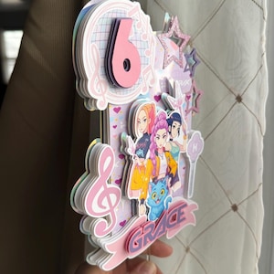 May include: A layered paper cake topper with a pink number 6, musical notes, and cartoon characters. The name "GRACE" is displayed on a pink banner. The topper has a star and a cat.