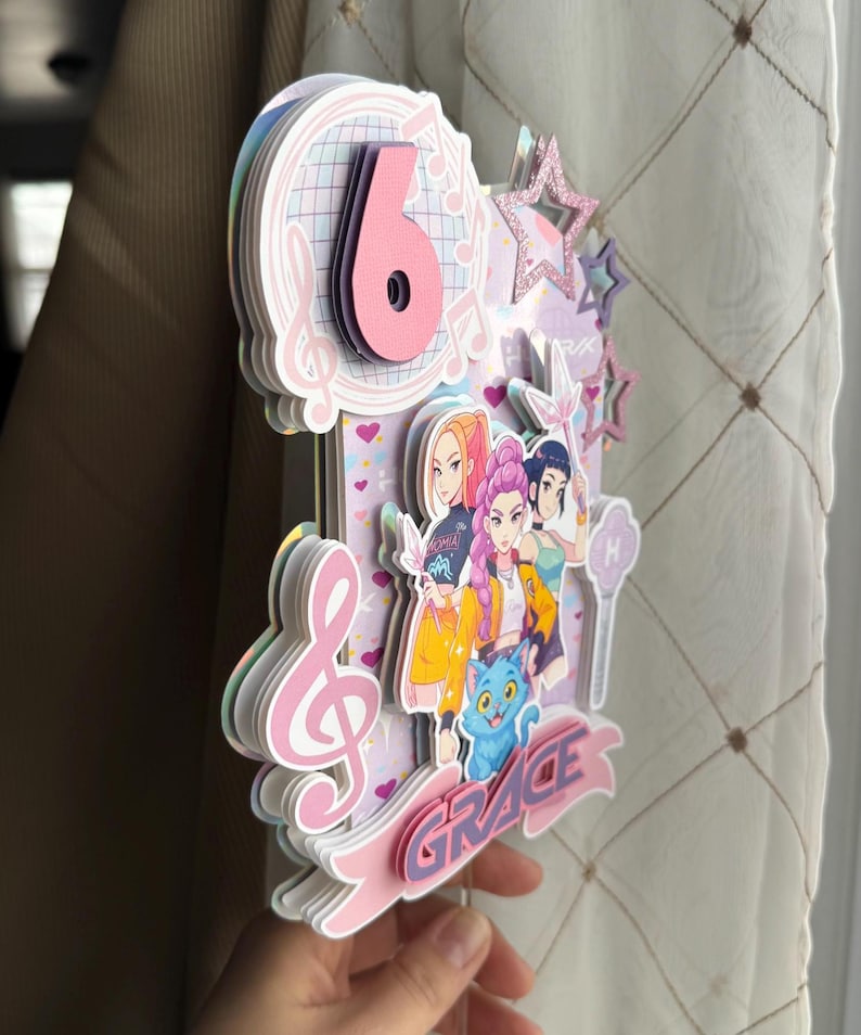 May include: A layered paper cake topper with a pink number 6, musical notes, and cartoon characters. The name "GRACE" is displayed on a pink banner. The topper has a star and a cat.