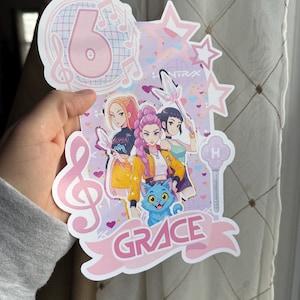 May include: A pink and white birthday cake topper with the number 6, musical notes, and stars. The topper features a cartoon illustration of four girls and a blue cat, with the name "GRACE" in a banner.
