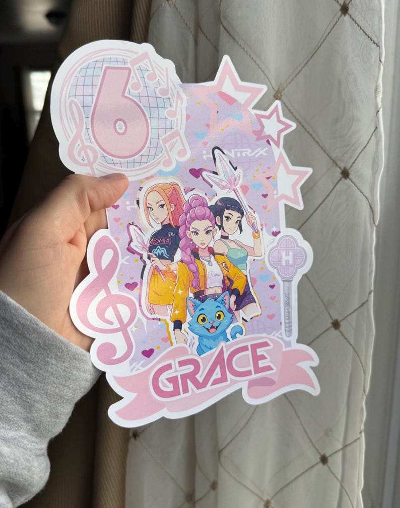 May include: A pink and white birthday cake topper with the number 6, musical notes, and stars. The topper features a cartoon illustration of four girls and a blue cat, with the name "GRACE" in a banner.