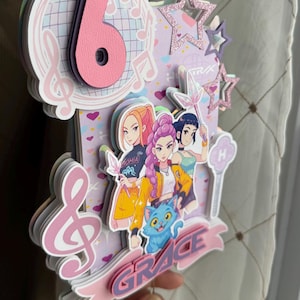 May include: A layered birthday cake topper with a musical theme. The design features a large pink number 6, musical notes, and illustrations of anime-style characters. The word "GRACE" is displayed on a banner, and there are glittery star accents.