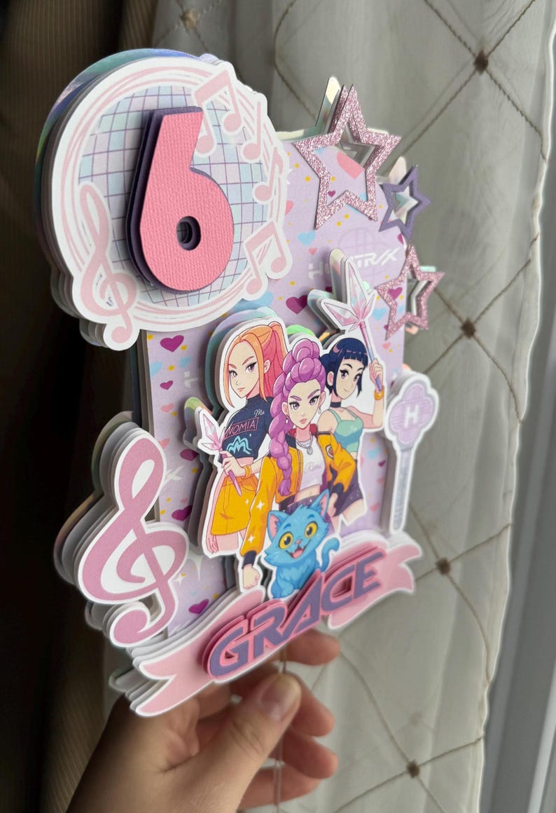 May include: A layered birthday cake topper with a musical theme. The design features a large pink number 6, musical notes, and illustrations of anime-style characters. The word "GRACE" is displayed on a banner, and there are glittery star accents.