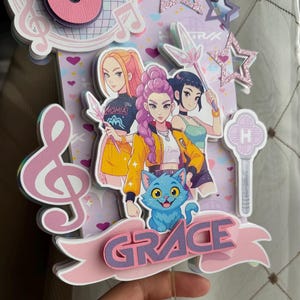 May include: A layered cake topper featuring three anime-style girls, a blue cat, and the name "GRACE" on a pink banner. The design includes musical notes, stars, and a large number six.