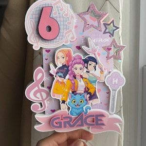May include: A colorful cake topper featuring a pink number 6, musical notes, and three cartoon girls. The topper includes a blue cat, the name "GRACE", and star accents. A hand is holding the topper.