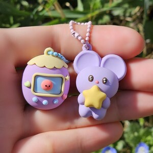 May include: Two purple keychains. One is shaped like a retro handheld game with a pink face on the screen. The other is a mouse holding a yellow star. Both keychains are attached to a beaded chain.