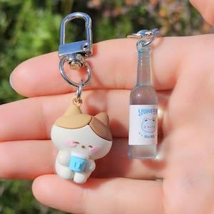 Pode incluir: Dois chaveiros: um apresenta um gato de desenho animado segurando uma xícara azul, o outro uma garrafa em miniatura com um rótulo. Ambos os chaveiros têm fechos e anéis prateados. O gato é branco e marrom, e a garrafa é transparente com um rótulo azul e branco.