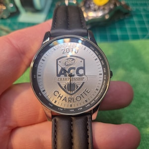 Op de afbeelding: Zilveren horloge met een zwarte leren band. De wijzerplaat toont de tekst "2010", "Dr. Pepper", "ACC CHAMPIONSHIP" en "CHARLOTTE". Het horloge heeft een zilveren rand met zwarte markeringen.