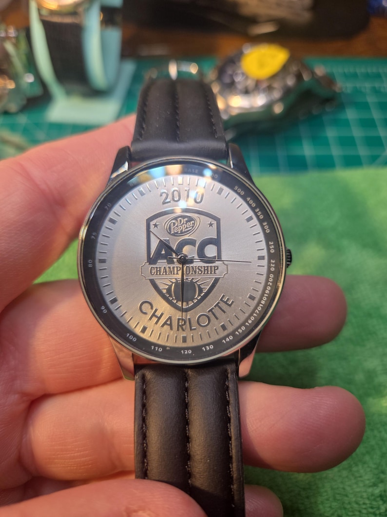 Op de afbeelding: Zilveren horloge met een zwarte leren band. De wijzerplaat toont de tekst "2010", "Dr. Pepper", "ACC CHAMPIONSHIP" en "CHARLOTTE". Het horloge heeft een zilveren rand met zwarte markeringen.