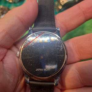 Op de afbeelding: Een zilverkleurig polshorloge met een zwarte leren band. De wijzerplaat van het horloge is bekrast en de achterkant van het horloge heeft de tekst "JAPAN MOVT". Het horloge wordt door een persoon vastgehouden.