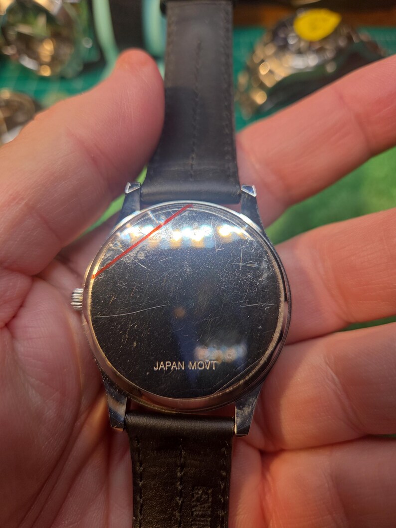 Op de afbeelding: Een zilverkleurig polshorloge met een zwarte leren band. De wijzerplaat van het horloge is bekrast en de achterkant van het horloge heeft de tekst "JAPAN MOVT". Het horloge wordt door een persoon vastgehouden.