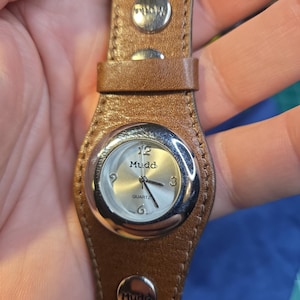 Könnte beinhalten: Braune Mudd-Quarzuhr aus Leder mit silbernem Zifferblatt und Edelstahl-Lünette. Die Uhr hat ein braunes Lederarmband mit silbernen Nieten und dem Mudd-Logo. Das Zifferblatt zeigt die Zeit mit silbernen Zeigern und Ziffern an.