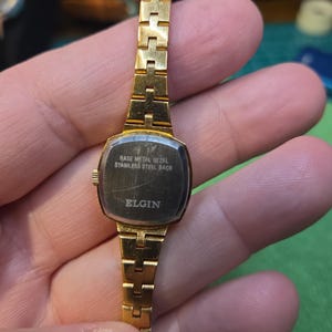 Peut inclure: Une montre Elgin dor&eacute;e avec un cadran rectangulaire et un bracelet dor&eacute; assorti. Le cadran de la montre est sombre avec le texte "BASE METAL BEZEL STAINLESS STEEL BACK ELGIN". La montre est tenue dans une main.