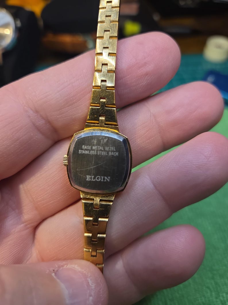 Peut inclure: Une montre Elgin dor&eacute;e avec un cadran rectangulaire et un bracelet dor&eacute; assorti. Le cadran de la montre est sombre avec le texte "BASE METAL BEZEL STAINLESS STEEL BACK ELGIN". La montre est tenue dans une main.
