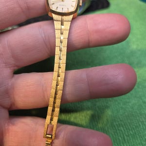 Peut inclure: Une montre-bracelet Elgin Quartz vintage. La montre dor&eacute;e a un cadran rectangulaire et un bracelet assorti. Le cadran est blanc avec des index et des aiguilles noirs. Le bracelet a un design textur&eacute; et segment&eacute;.