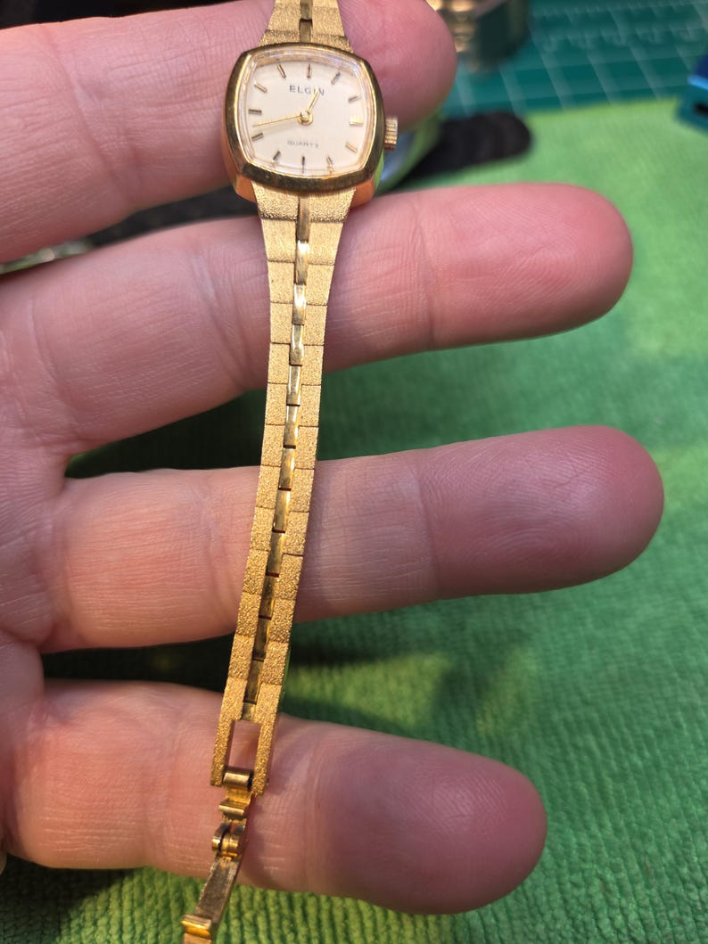 Peut inclure: Une montre-bracelet Elgin Quartz vintage. La montre dor&eacute;e a un cadran rectangulaire et un bracelet assorti. Le cadran est blanc avec des index et des aiguilles noirs. Le bracelet a un design textur&eacute; et segment&eacute;.