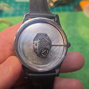 Op de afbeelding: Een horloge met een zwarte leren band, de achterkant is open en toont het interne mechanisme. De wijzerplaat is zilverkleurig, met een batterij en printplaat zichtbaar. Het horloge wordt vastgehouden, waardoor de interne componenten zichtbaar zijn.