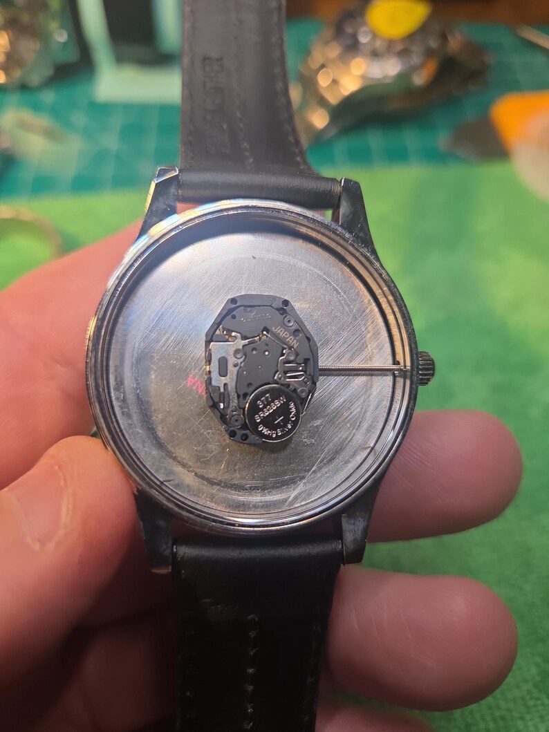 Op de afbeelding: Een horloge met een zwarte leren band, de achterkant is open en toont het interne mechanisme. De wijzerplaat is zilverkleurig, met een batterij en printplaat zichtbaar. Het horloge wordt vastgehouden, waardoor de interne componenten zichtbaar zijn.