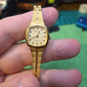 Peut inclure: Une montre-bracelet Elgin Quartz vintage. Le bo&icirc;tier dor&eacute; de la montre est rectangulaire, avec un bracelet assorti. Le cadran cr&egrave;me affiche des index et des aiguilles noirs. Le bracelet est textur&eacute; et &agrave; maillons.