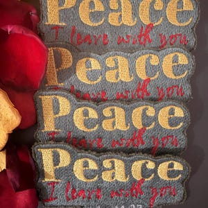May include: Four embroidered patches with the word "Peace" in gold thread, the phrase "I leave with you" in red, and the Bible verse John 14:27. The patches are gray with a decorative border. Red and yellow flower petals surround the patches.