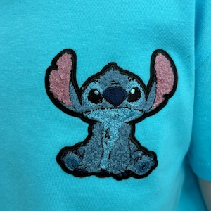 Puede incluir: Una camiseta azul claro con una imagen bordada del personaje Stitch de la pel&iacute;cula Lilo & Stitch. La imagen de Stitch es azul con orejas internas rosas y un contorno negro. La camiseta es de color azul claro liso.