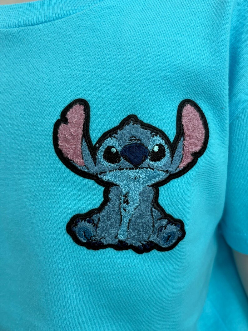 Puede incluir: Una camiseta azul claro con una imagen bordada del personaje Stitch de la pel&iacute;cula Lilo & Stitch. La imagen de Stitch es azul con orejas internas rosas y un contorno negro. La camiseta es de color azul claro liso.