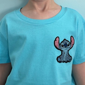 Puede incluir: Una camiseta azul claro con un peque&ntilde;o parche bordado del personaje Stitch. El parche es azul con orejas rosas y un contorno negro. La camiseta tiene mangas cortas y cuello redondo. La camiseta es de material suave.