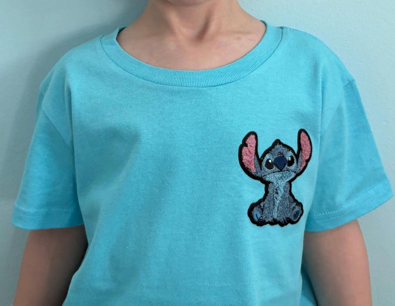 Puede incluir: Una camiseta azul claro con un peque&ntilde;o parche bordado del personaje Stitch. El parche es azul con orejas rosas y un contorno negro. La camiseta tiene mangas cortas y cuello redondo. La camiseta es de material suave.