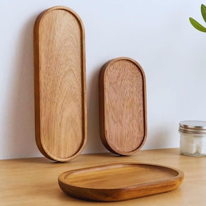 Può includere: Tre vassoi ovali in legno di diverse dimensioni. I vassoi sono in legno marrone chiaro con venature visibili. Un vassoio è montato su una parete bianca, mentre gli altri poggiano su una superficie di legno. I vassoi hanno un bordo rialzato.