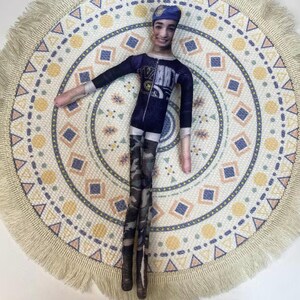 May include: A handmade doll with a printed face, wearing a navy blue jumpsuit with a graphic design and camouflage leggings. The doll has a blue cap and is lying on a round, patterned rug with a fringed edge. The rug features geometric shapes in various colors.