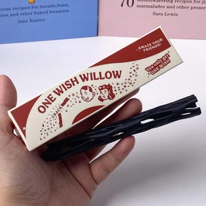May include: A rectangular box labeled ONE WISH WILLOW with red and cream graphics, featuring illustrations of two faces and the text AMAZE YOUR FRIENDS and You only get ONE WISH. Beside the box is a long, black, cylindrical object with cutouts.