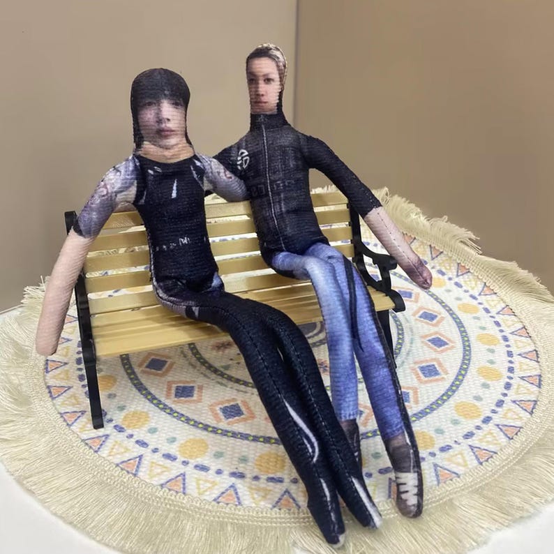 May include: Two fabric dolls, each with a printed face, sit on a miniature wooden bench. One doll wears a black outfit with a tattoo-like design on the arm, while the other wears a black jacket and blue jeans. They are posed on a patterned mat.