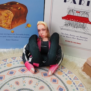 Peut inclure: Une figurine ressemblant &agrave; une poup&eacute;e avec un visage imprim&eacute; et des v&ecirc;tements noirs avec des accents roses. La figurine est assise sur un tapis &agrave; motifs, avec un livre intitul&eacute; "PARIS" et un autre livre sur la p&acirc;tisserie en arri&egrave;re-plan.