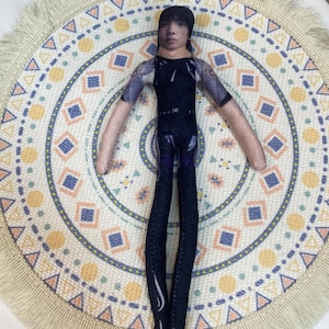 May include: A handmade doll with a printed face, wearing a black and purple outfit. The doll has a black cap and long, slender limbs. It is positioned on a circular, patterned fabric with a cream-colored fringe.