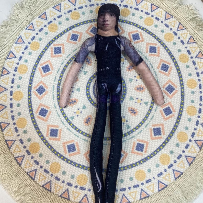 May include: A handmade doll with a printed face, wearing a black and purple outfit. The doll has a black cap and long, slender limbs. It is positioned on a circular, patterned fabric with a cream-colored fringe.