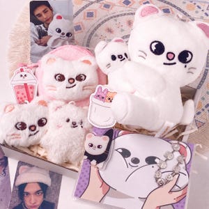 May include: A collection of white plush cat-themed merchandise including a large plush toy, several smaller plush charms, a pink plush pouch, a metal chain bracelet with cat charms, and two photographic cards featuring a person holding a plush cat.