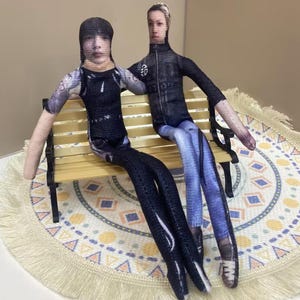 May include: Two fabric dolls, each with a printed face, sit on a miniature wooden bench. One doll wears a black outfit, the other a black jacket and blue jeans. The dolls are on a patterned surface.