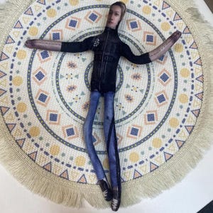 May include: A fabric doll with a realistic face, wearing a black jacket, blue jeans, and black shoes. The doll is lying on a round, cream-colored rug with a geometric pattern in blue, yellow, and orange, and a fringed edge.