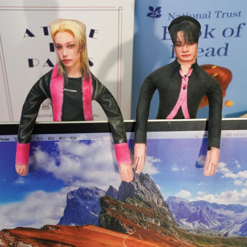 Peut inclure: Deux figurines d&eacute;coratives, chacune avec un visage et des bras humains, sont pos&eacute;es sur un &eacute;cran d'ordinateur portable. L'une a les cheveux blonds et une tenue rose et noire. L'autre a les cheveux fonc&eacute;s et une tenue noire avec des touches de rose. L'&eacute;cran affiche un paysage de montagne.