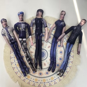 May include: Five handmade fabric dolls with printed faces and clothing. The dolls are arranged on a round, beige, patterned mat. Each doll has a unique outfit, including jackets, shirts, and trousers in shades of blue, black, and white.