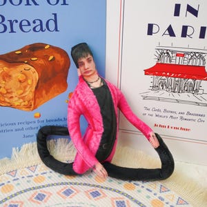 Peut inclure: Une poup&eacute;e en tissu faite &agrave; la main en position de yoga, v&ecirc;tue d'une veste rose sur un haut noir et un pantalon noir. La poup&eacute;e est plac&eacute;e devant deux couvertures de livres, l'une avec une illustration de pain et l'autre avec une sc&egrave;ne de caf&eacute; parisien.