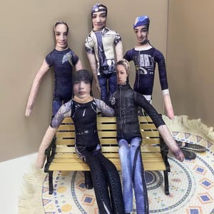 May include: A group of five fabric dolls, each with a printed face and wearing different outfits. The dolls are posed in a group, some standing and some seated on a small bench. The outfits include jeans, jackets, and t-shirts.