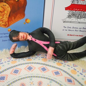 Peut inclure: Une poup&eacute;e v&ecirc;tue d'une chemise noire &agrave; manches longues avec des bretelles roses et un pantalon noir, allong&eacute;e sur un tapis &agrave; motifs. La poup&eacute;e est devant un livre avec le texte "recipes and othe" et un autre livre avec le texte "THE CAF&Eacute;S, BISTROS, AND BRAS OF THE WORLD'S MOST ROMANTIC".