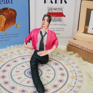 Peut inclure: Une figurine de personne portant un blazer rose, une chemise blanche, une cravate noire et un pantalon noir. La figurine est pos&eacute;e sur un tapis rond &agrave; motifs. Des livres et un cadre en bois sont en arri&egrave;re-plan.